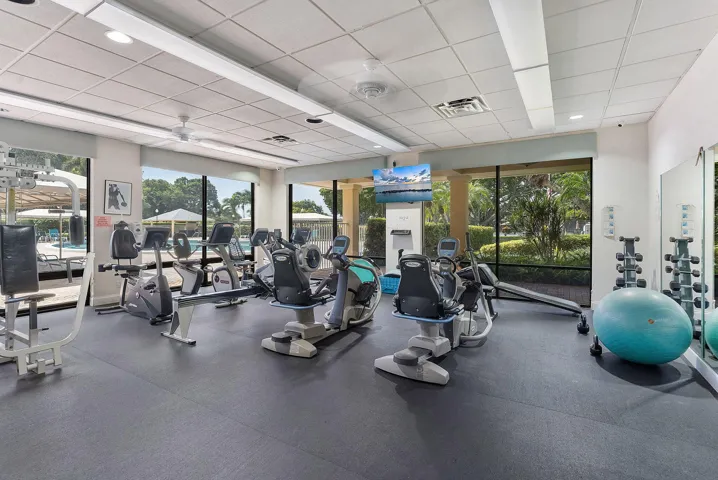State-of-the-art fitness facility featuring multiple cardio and strength training stations, durable rubberized flooring, abundant natural light from expansive glass walls and windows, and direct access to manicured landscaping and outdoor grounds