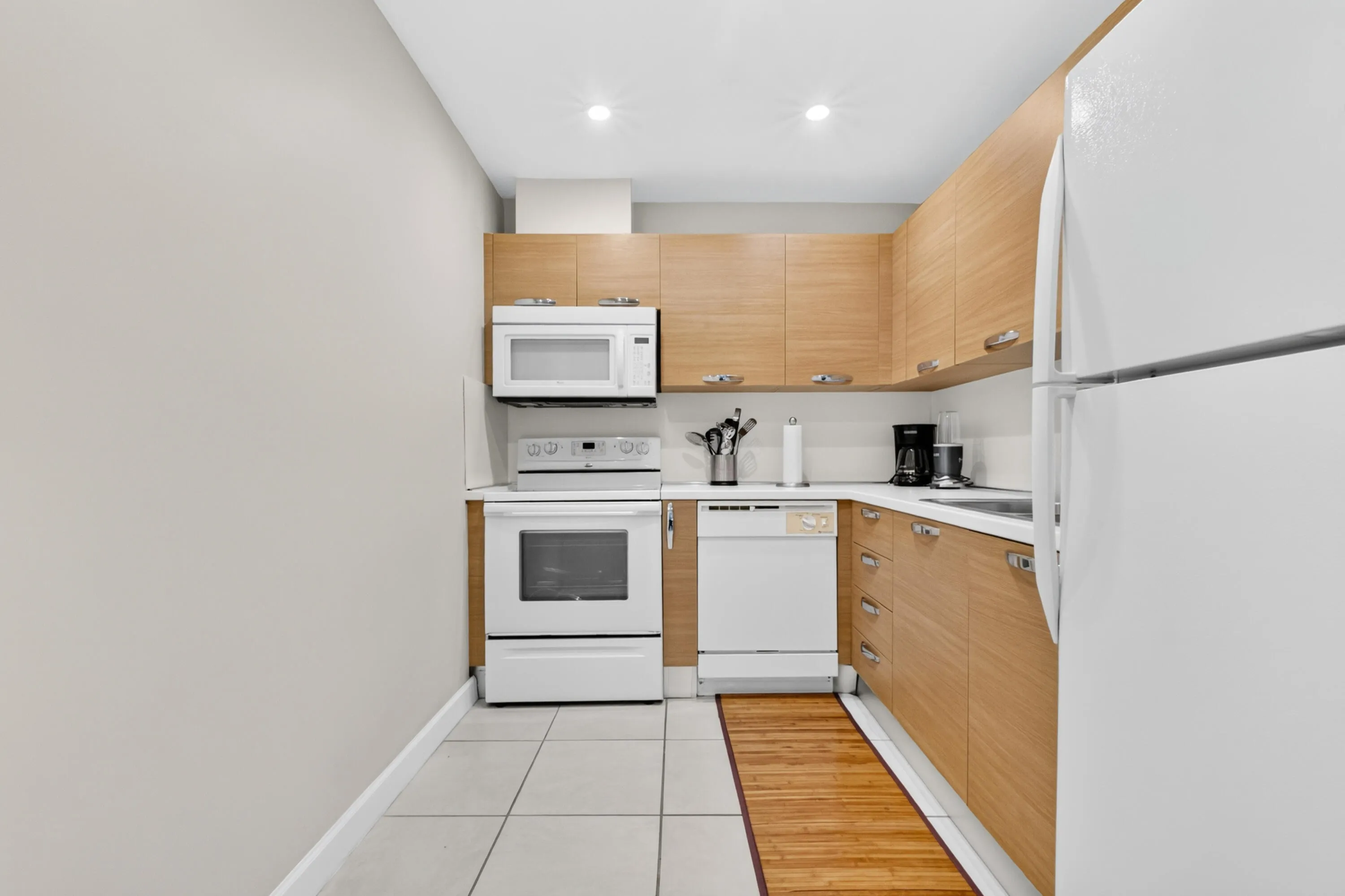 Galley kitchen featuring natural wood-finish cabinetry, white appliances including a gas range and dishwasher, tile flooring, and recessed ceiling lighting
