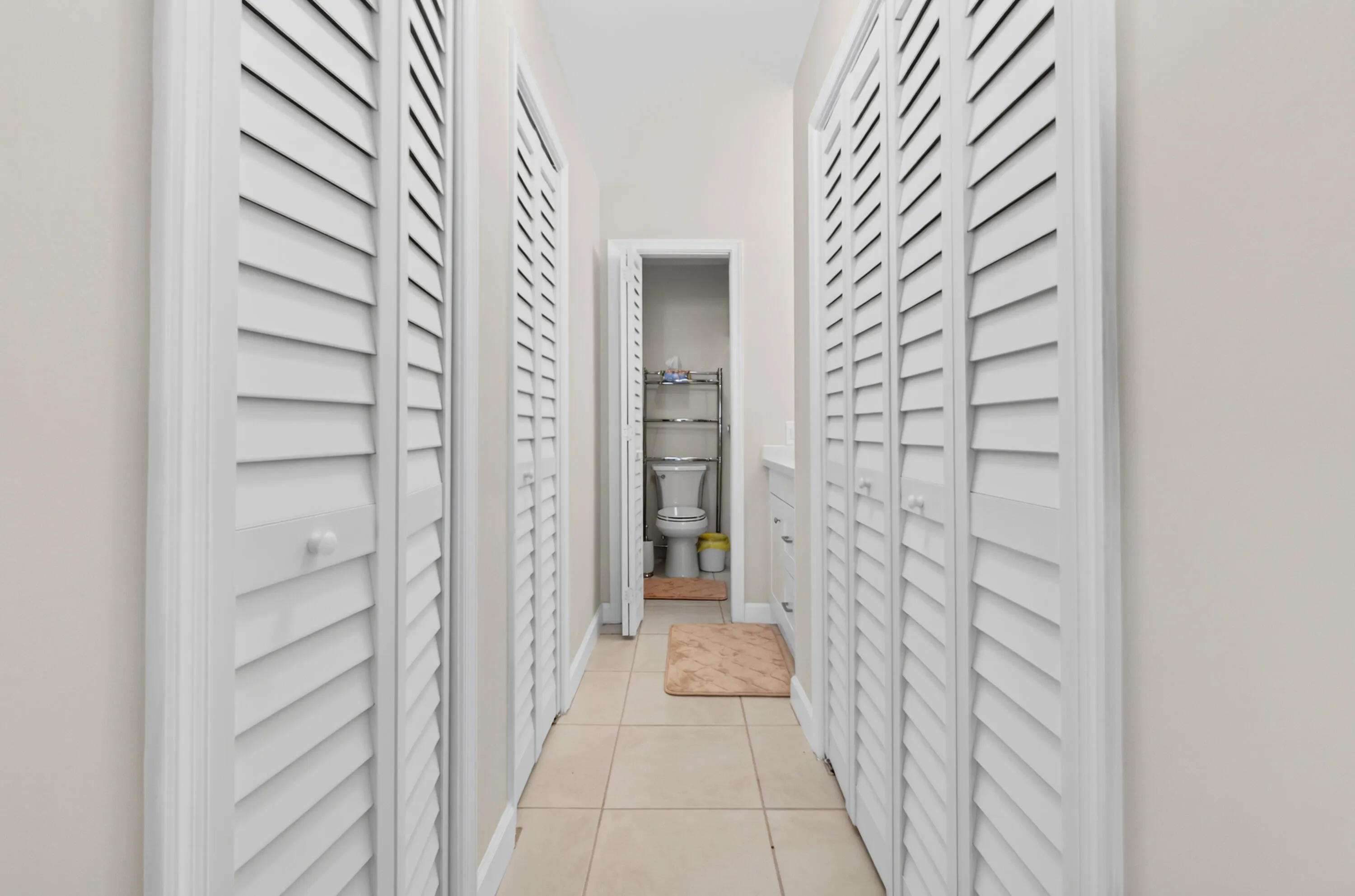 Hallway featuring white louvered closet doors on either side, neutral tile flooring, and bright overhead lighting creating a clean, organized passage to the adjacent bathroom