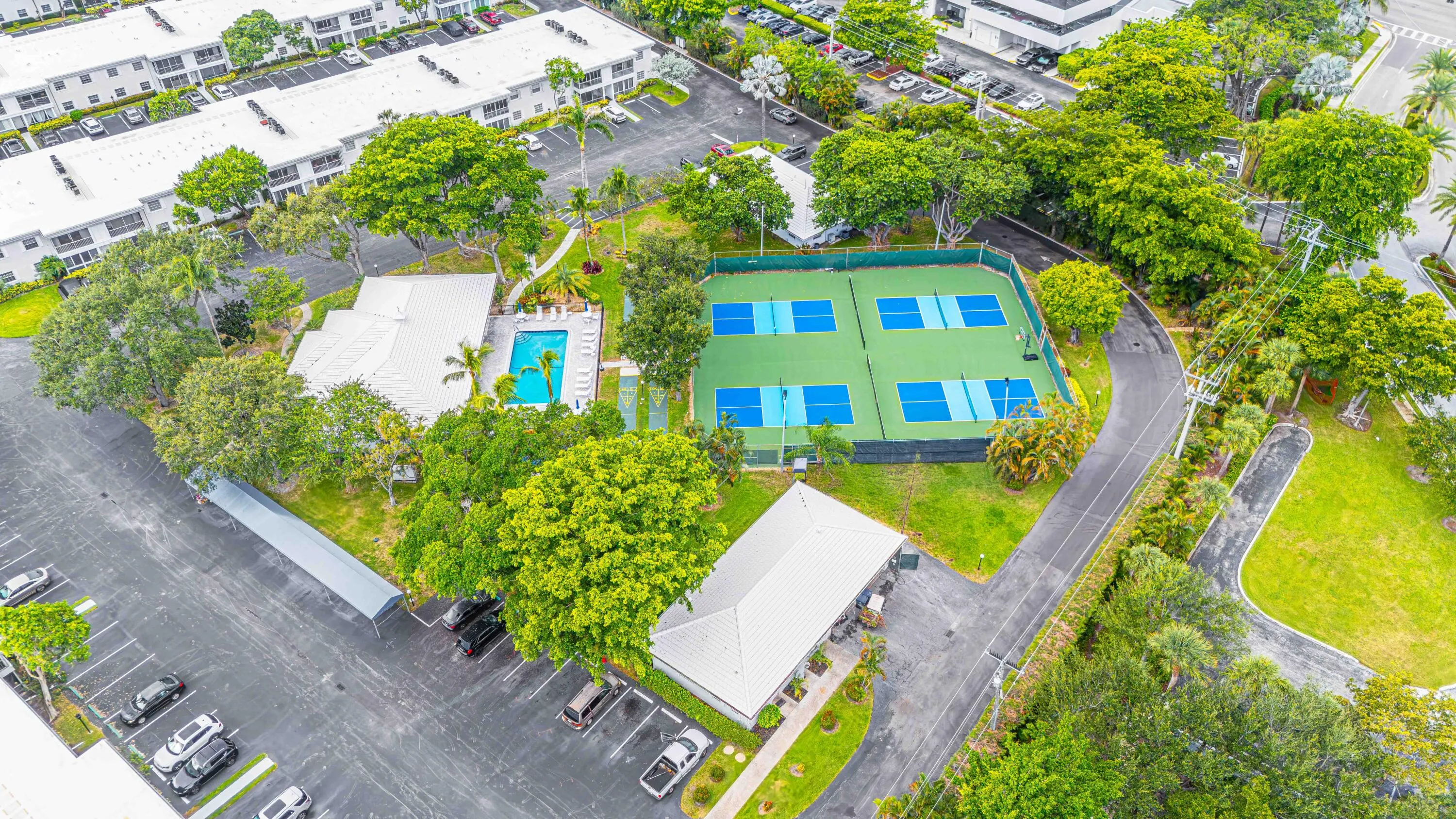 Aerial view of a recreational facility featuring a regulation-size tennis court with blue playing surface, adjacent lap pool with defined lanes, mature landscaping throughout the grounds, and ample parking