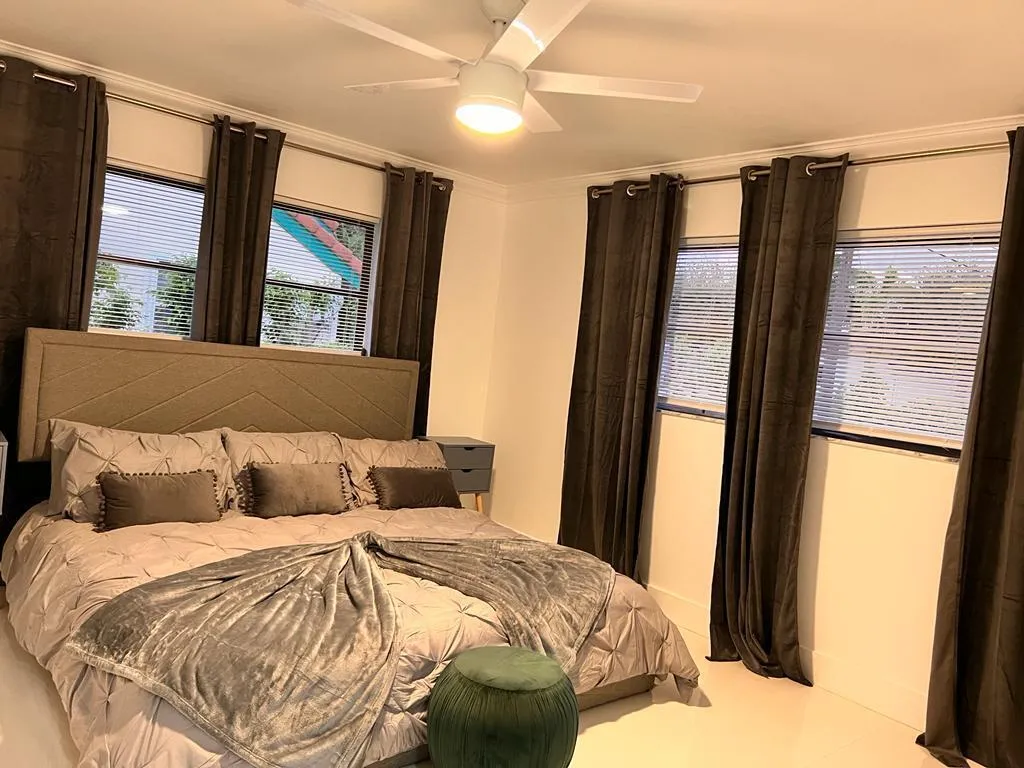 Spacious primary bedroom featuring multiple windows with blackout drapery, ceiling fan with integrated lighting, wood-finish flooring, and a platform bed with upholstered headboard and coordinating bedding ensemble