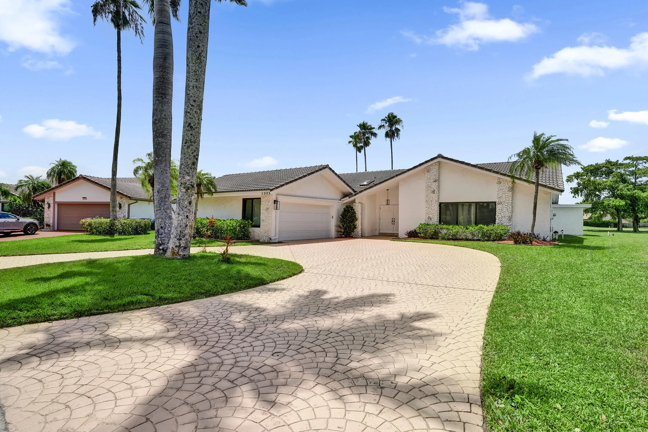 Paved circular driveway, two attached garages with roll-up doors, mature palm trees, stucco exterior, and a tiled roof