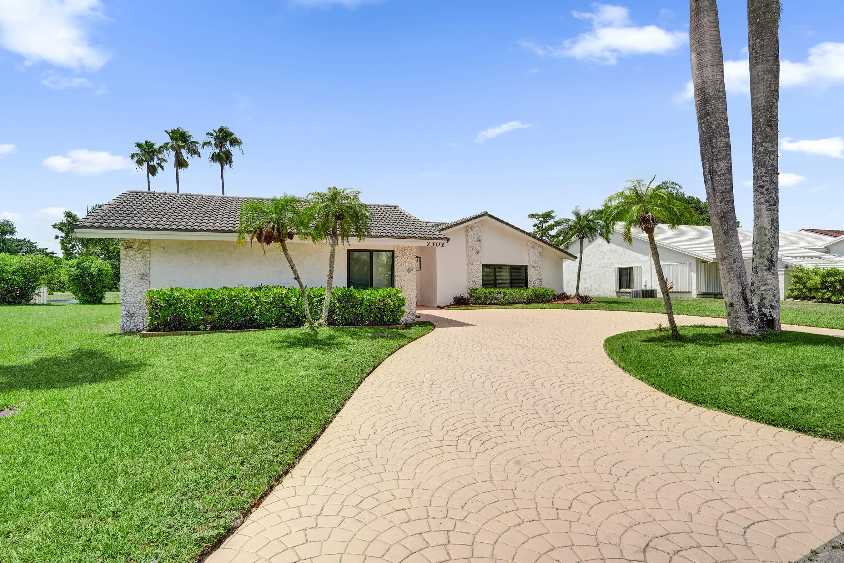 White stucco exterior with a tiled roof, curved paver driveway, manicured lawn, and mature palm trees