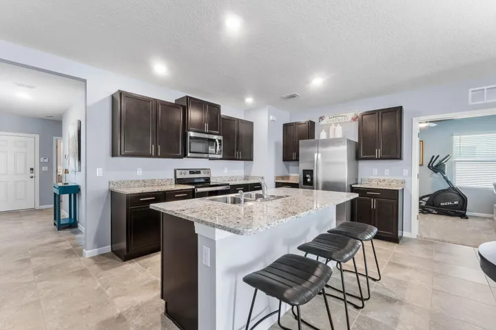 Spacious kitchen featuring dark wood cabinetry, stainless steel appliances, a large center island with a double basin sink, light-toned stone countertops, and tile flooring