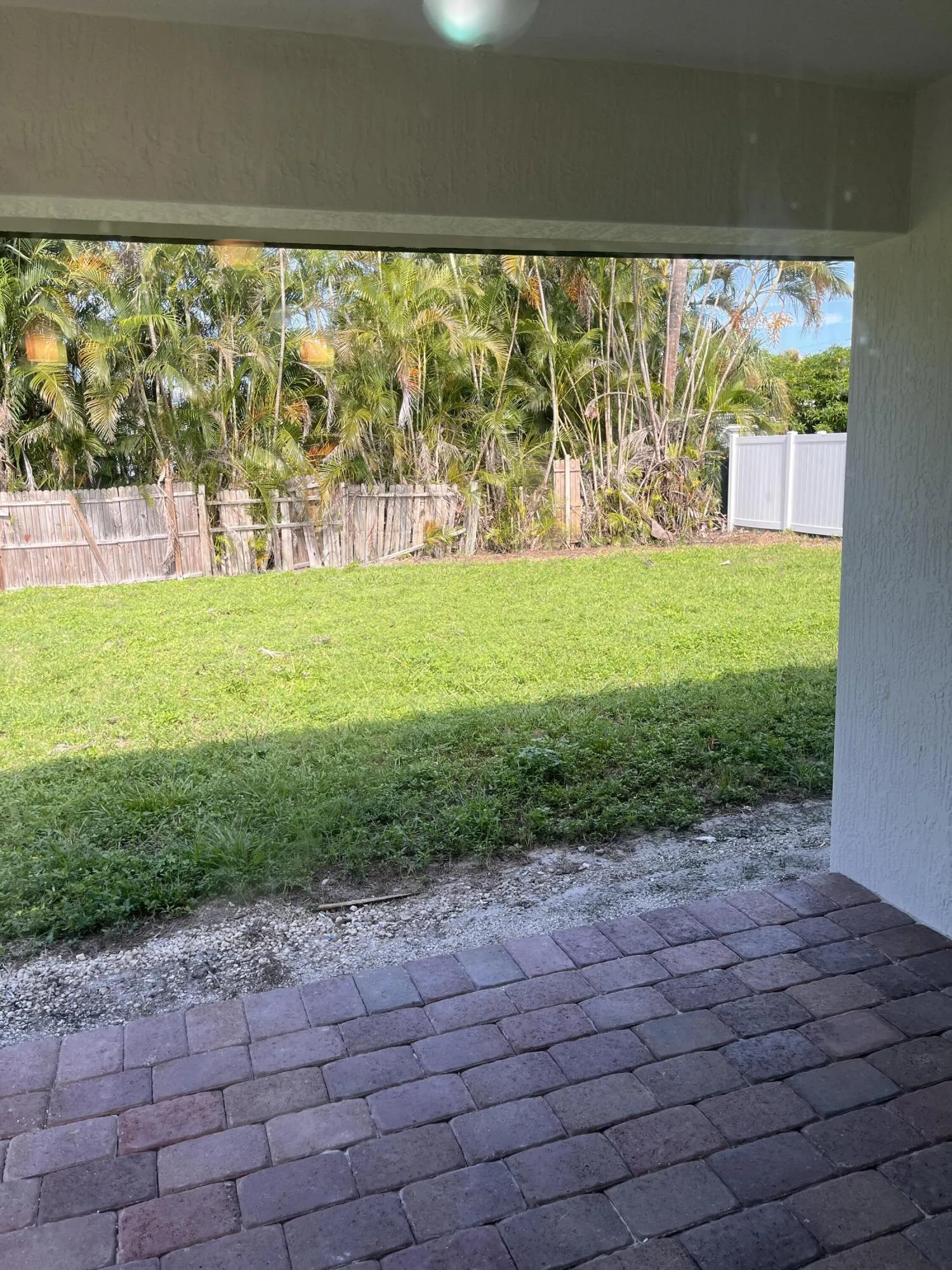 Brick paver patio with a grassy yard, surrounded by wood fencing and mature tropical foliage