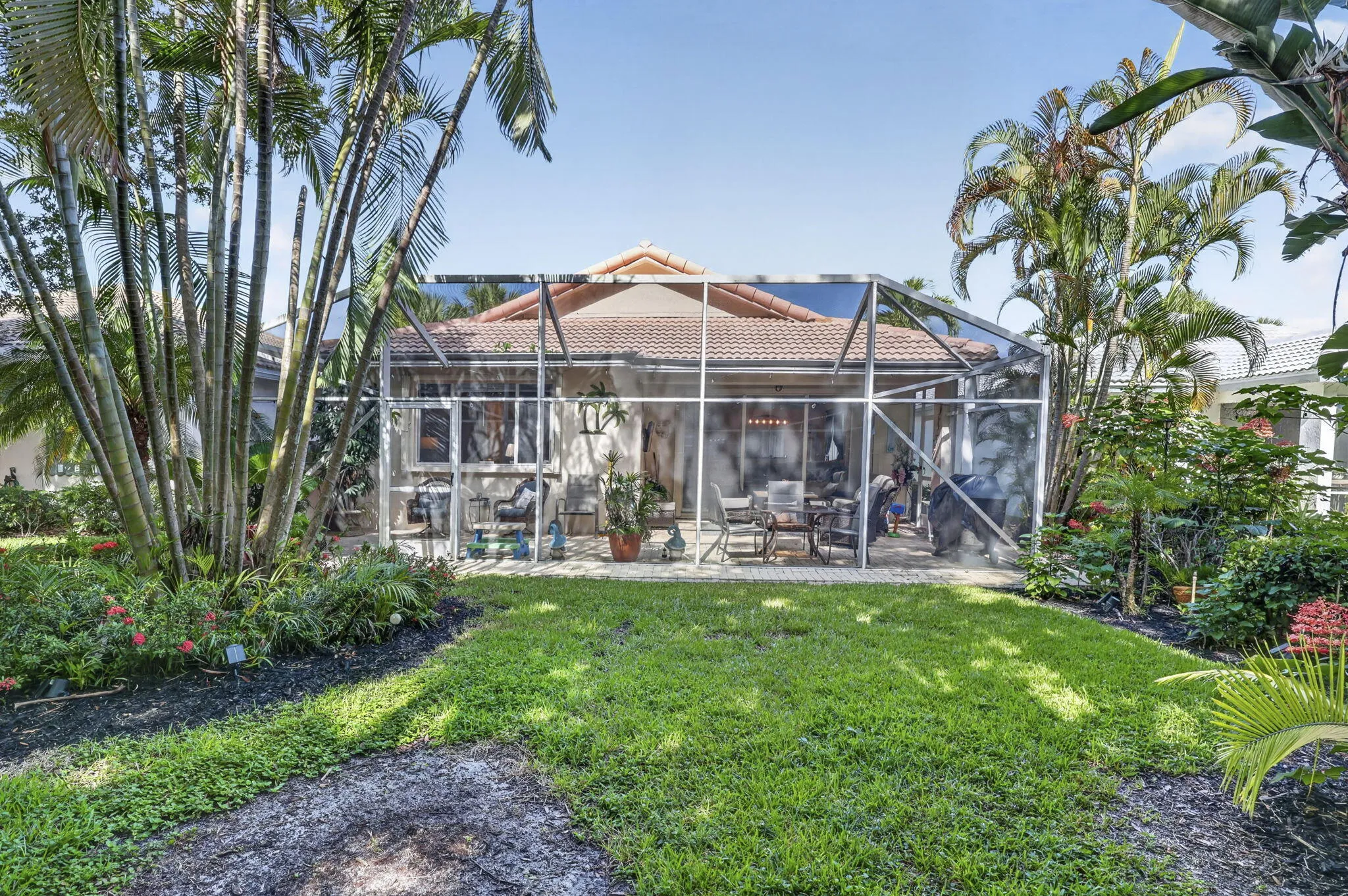 Spacious backyard oasis featuring a manicured lawn, mature tropical landscaping with ornamental palms, and a screened-in pool enclosure with a tile roof