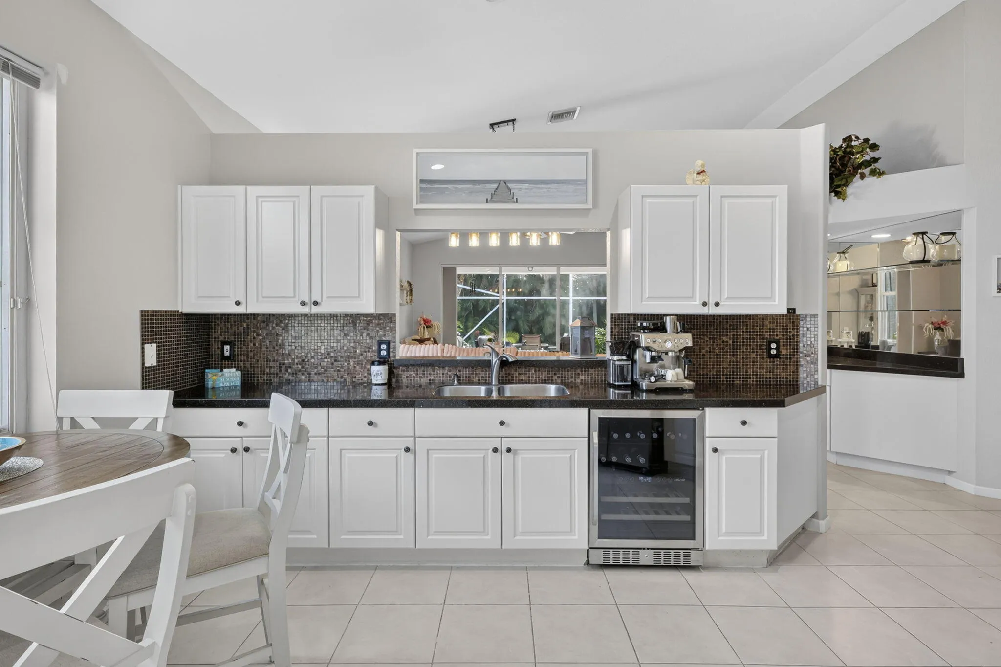 White cabinetry with dark hardware, dark stone countertops, mosaic tile backsplash, integrated beverage cooler, and a double basin stainless steel sink