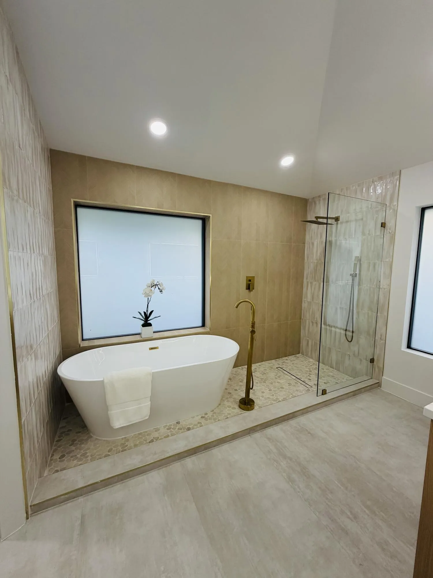 Freestanding soaking tub with floor-mounted brass-finish filler, glass-enclosed shower with brass-finish fixtures, and light-toned wall and floor tiling