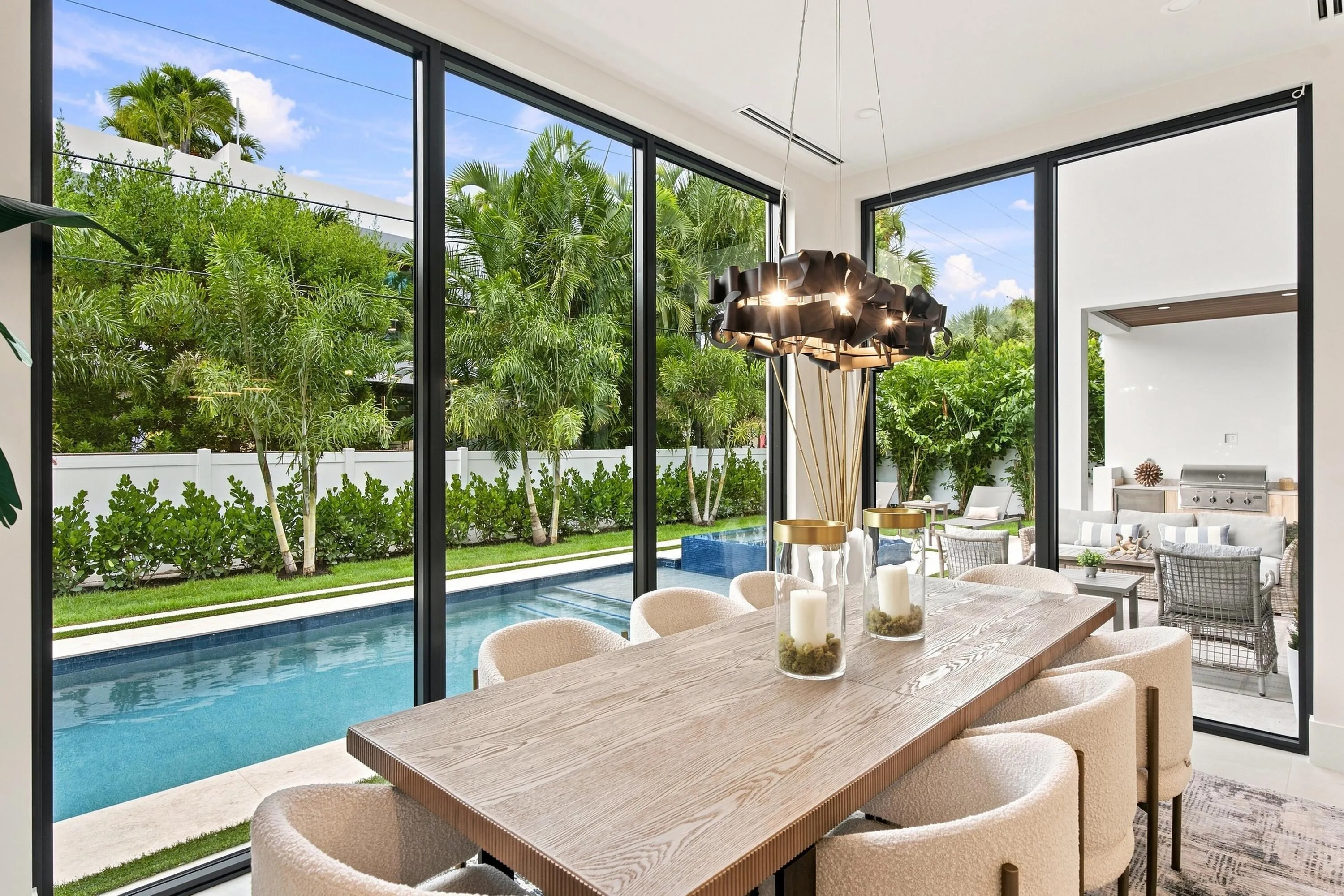 Expansive sliding glass doors, contemporary chandelier, wood-finish dining table, recessed lighting, and a private pool