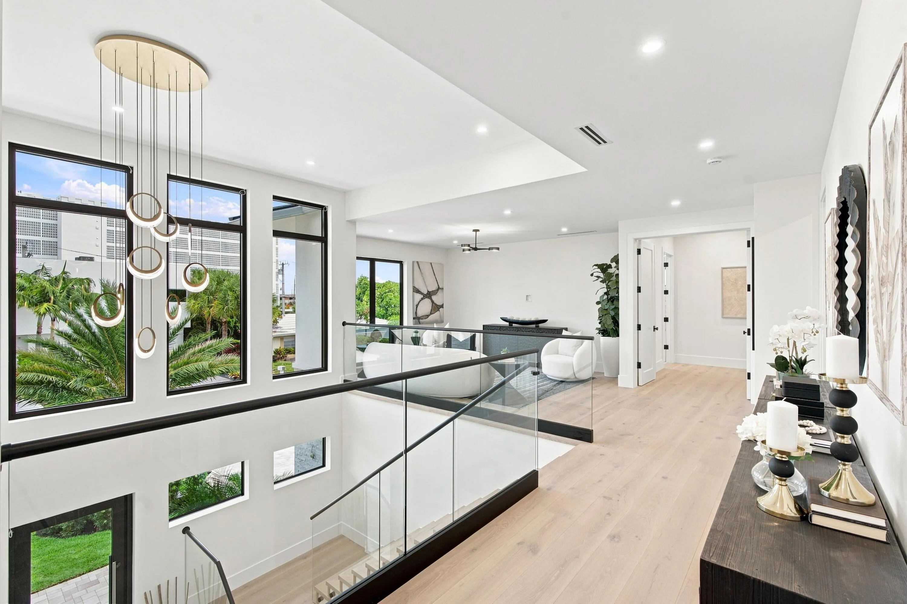 Contemporary chandelier with multiple rings, glass railing system, light wood-finish flooring, recessed ceiling lighting, and black-framed windows