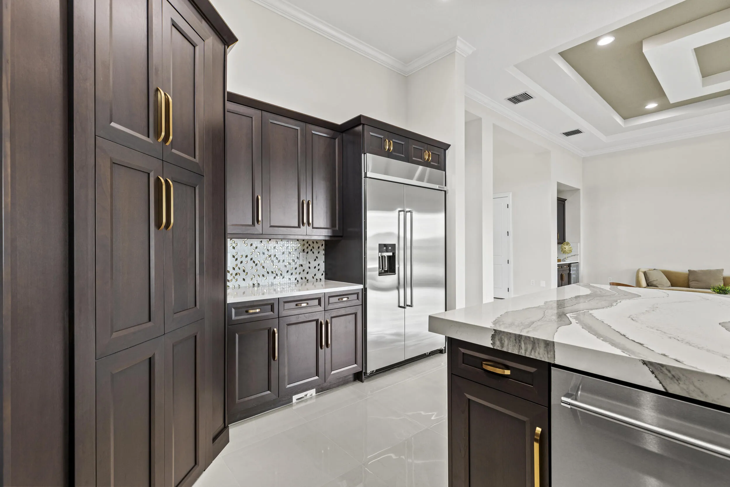 Dark wood cabinetry with gold-tone hardware, stainless steel refrigerator, patterned tile backsplash, light-toned flooring, and a tray ceiling