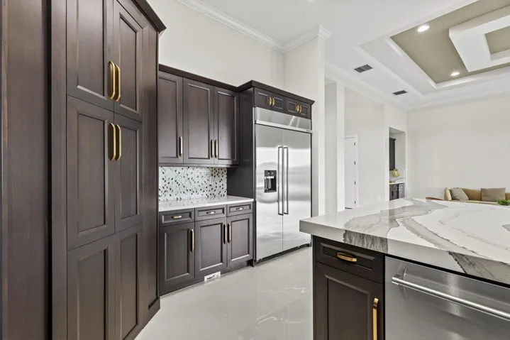Dark wood cabinetry with gold-tone hardware, stainless steel refrigerator, patterned tile backsplash, light-toned flooring, and a tray ceiling
