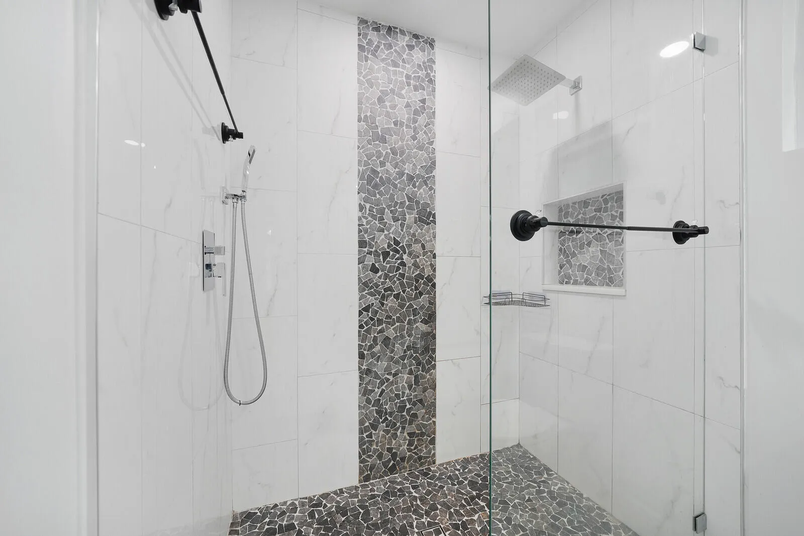 Glass-enclosed shower with pebble-finish flooring and accent wall, white tile surround, rain shower head, and built-in shower niche