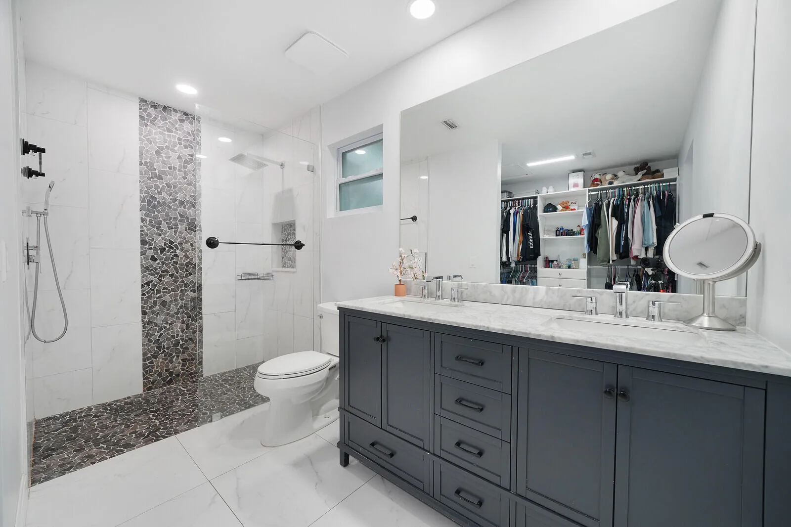 Walk-in shower with pebble tile flooring and an accent wall, dual vanity with a light-toned countertop, integrated rectangular sinks, and a large wall-mounted mirror