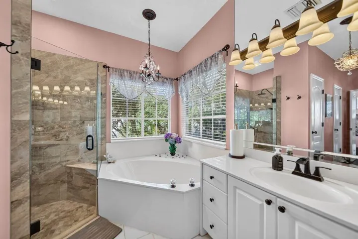 Tiled walk-in shower with a glass enclosure, corner soaking tub, dual-sink vanity with white cabinetry, and a chandelier fixture