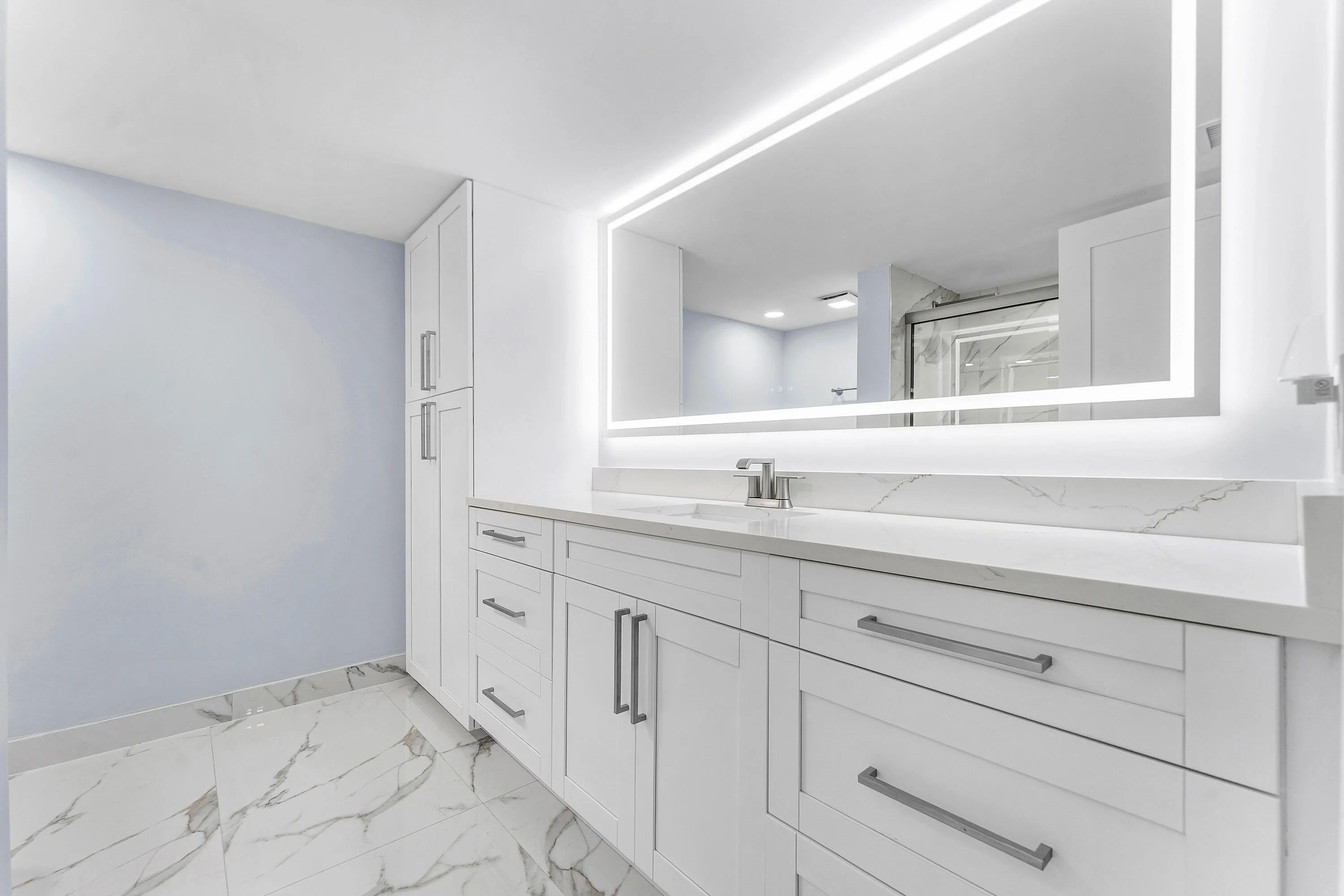 White shaker cabinetry with brushed nickel hardware, tile flooring, illuminated mirror, and a white countertop with integrated sink