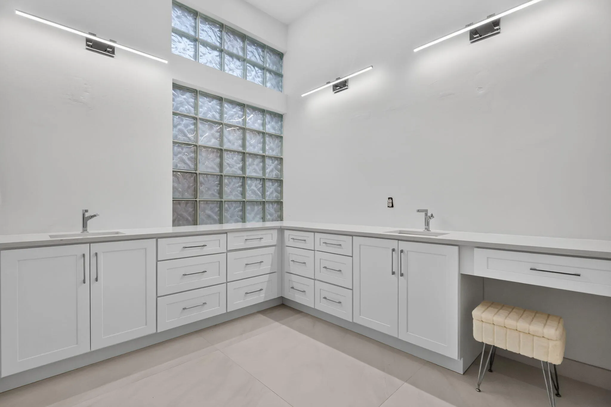 Dual sinks with modern faucets, extensive white cabinetry, light-toned countertops, contemporary wall sconces, and a block glass window