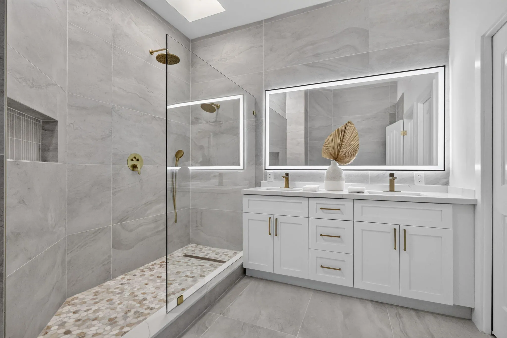 Dual vanity with white cabinetry and integrated sinks, illuminated mirror, walk-in shower with glass enclosure, gold-finish fixtures, and a skylight