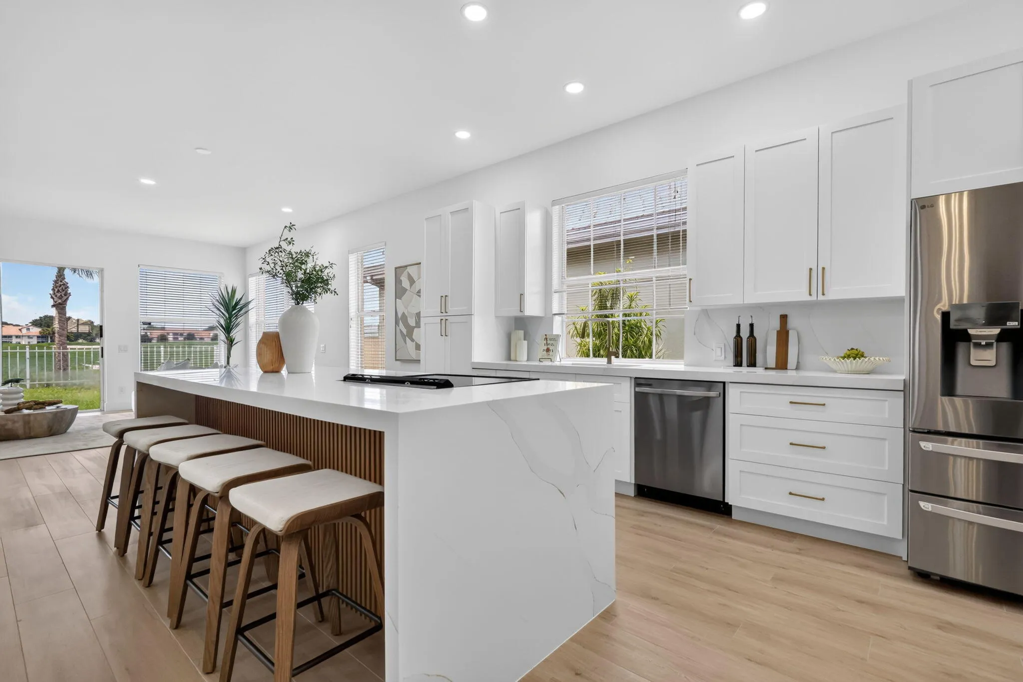 Modern kitchen featuring white cabinetry with integrated hardware, quartz waterfall island countertop, stainless steel appliances including dual refrigerators and dishwasher, wood-finish flooring, recessed ceiling lighting, and expansive windows overlooking outdoor space