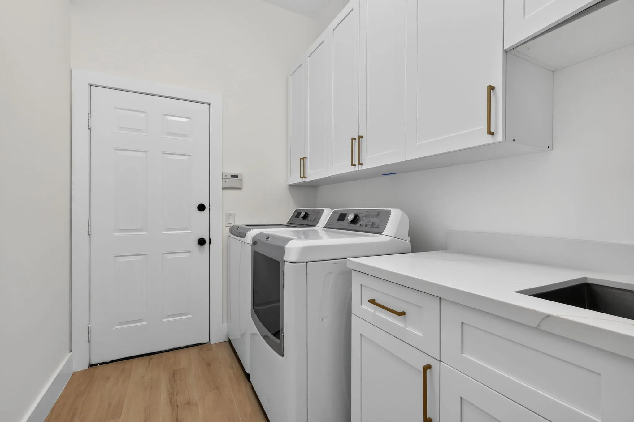 White shaker-style cabinetry with gold-tone hardware, a solid surface countertop with an integrated sink, wood-finish flooring, and crisp white walls