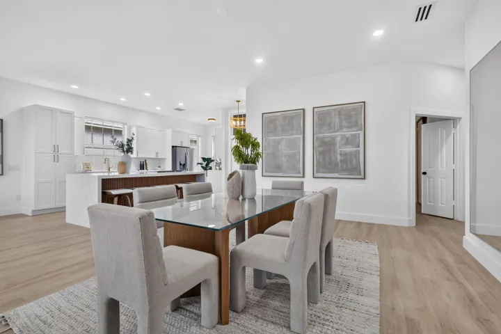 Open-concept dining and kitchen area featuring a wood-finish dining table with upholstered chairs, white cabinetry with integrated storage, stainless steel appliances, quartz countertops, recessed ceiling lighting, and wide-plank wood-finish flooring throughout