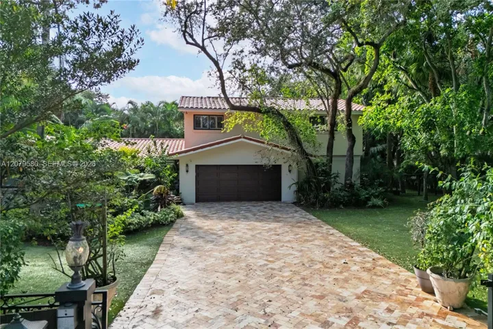 Two-story exterior featuring a clay tile roof, herringbone-pattern stone paver driveway, multi-car garage, and mature landscaping