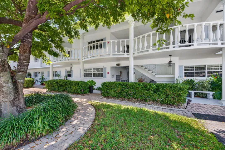 Multi-story building exterior featuring white cat walk, decorative columns, light-colored shutters, and mature landscaping
