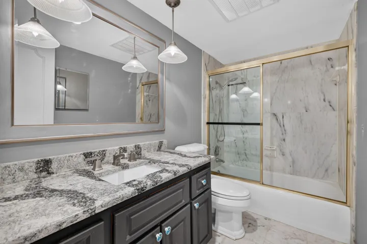 Granite countertop with integrated sink, dark wood-finish vanity, large framed mirror, glass-enclosed shower with tiled walls, and recessed ceiling lighting