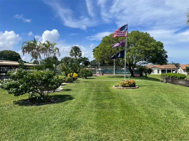 Expansive lawn featuring mature trees, manicured landscaping, a dedicated flagpole, and a fenced recreational court
