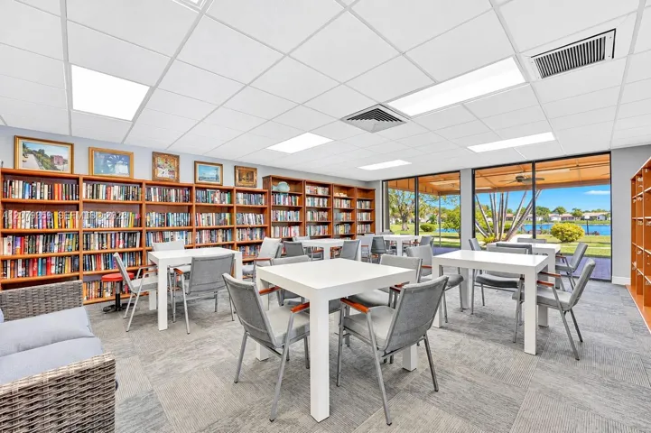 Expansive room featuring wall-to-wall built-in wooden bookshelves, modular square-grid acoustic ceiling tiles, recessed rectangular lighting, grey patterned carpeted flooring, and floor-to-ceiling glass panel windows