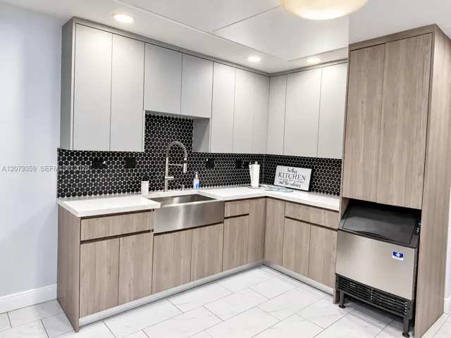 Custom cabinetry featuring two-tone wood-finish and matte-finish flat-panel doors, stainless steel farmhouse sink with gooseneck faucet, contrasting black penny-round tile backsplash, stone-finish countertops, and built-in under-counter ice machine