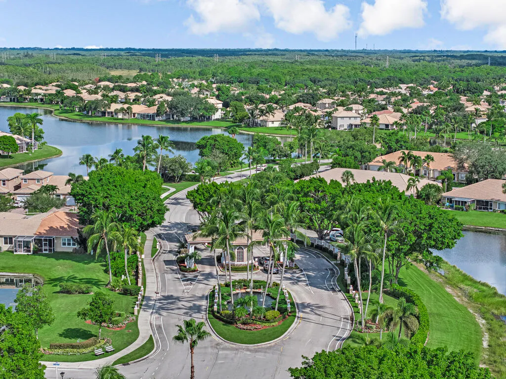 Lush tropical landscaping, mature palm trees, a gated community entrance, and homes with barrel tile roofs bordering a substantial lake