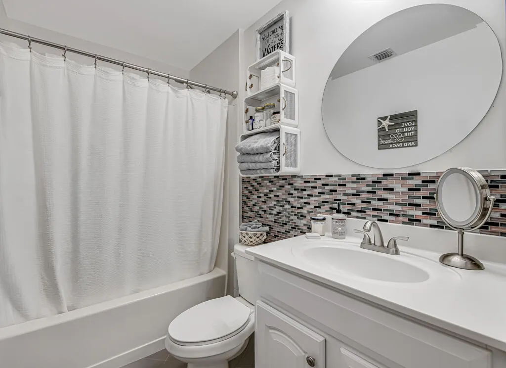 White vanity with integrated sink, mosaic tile backsplash, circular wall mirror, and a full-size tub with shower