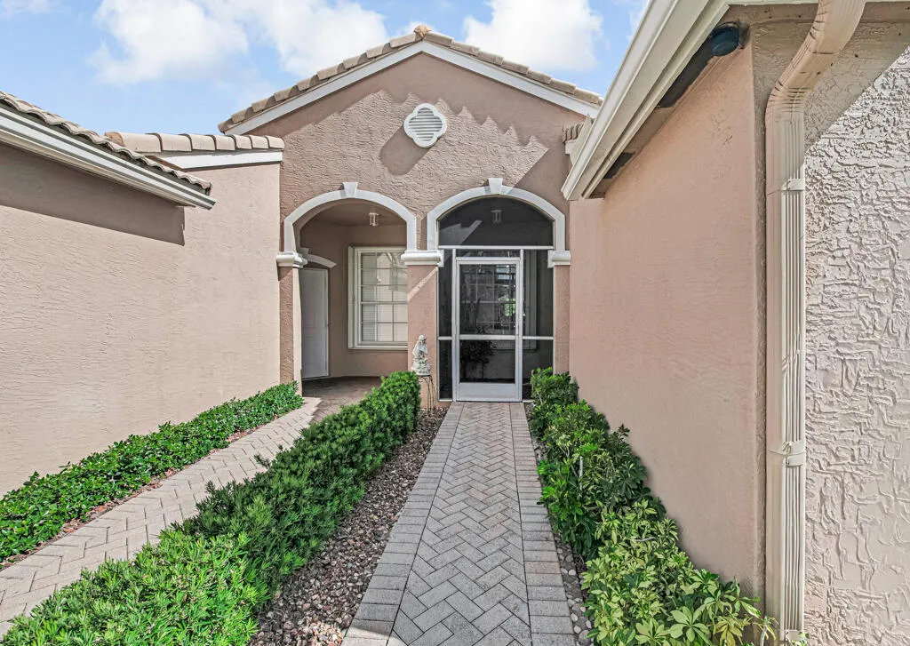 Paver walkway bordered by manicured shrubs, stucco exterior, arched entryways, and a screened front door