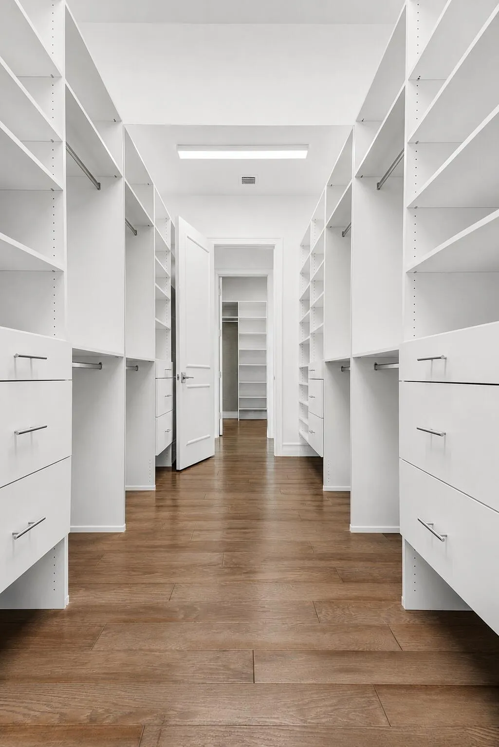 Spacious walk-in closet featuring extensive white shelving, built-in drawers with modern hardware, hanging rods, and wood-finish flooring