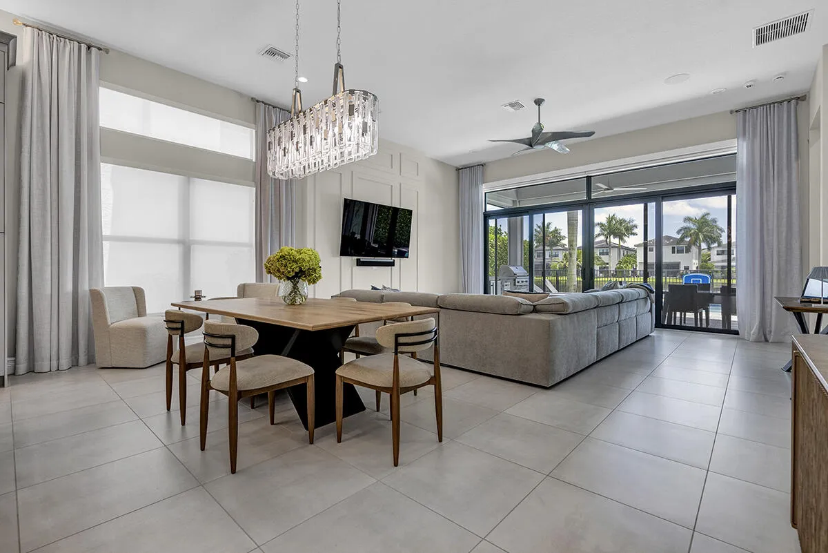 Spacious interior featuring large format tile flooring, a contemporary chandelier, a decorative accent wall, floor-to-ceiling windows with roller shades, and expansive sliding glass doors leading to an outdoor area