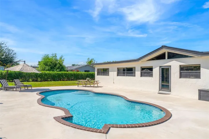 In-ground pool with masonry coping, expansive concrete patio, gabled roofline, and stucco exterior finish