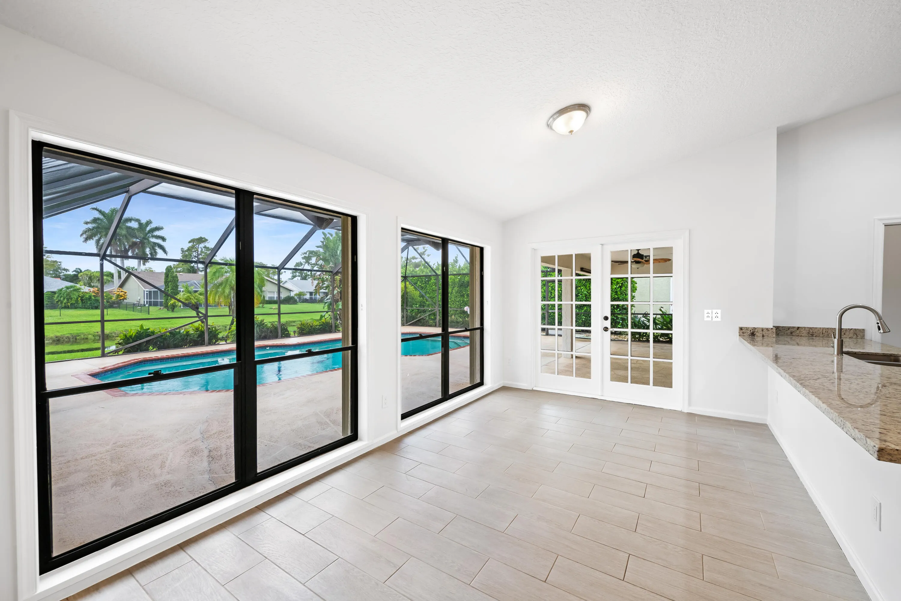 White vaulted ceiling, light-toned tile flooring, double French doors, large window overlooking a swimming pool, and a kitchen counter with an undermount sink