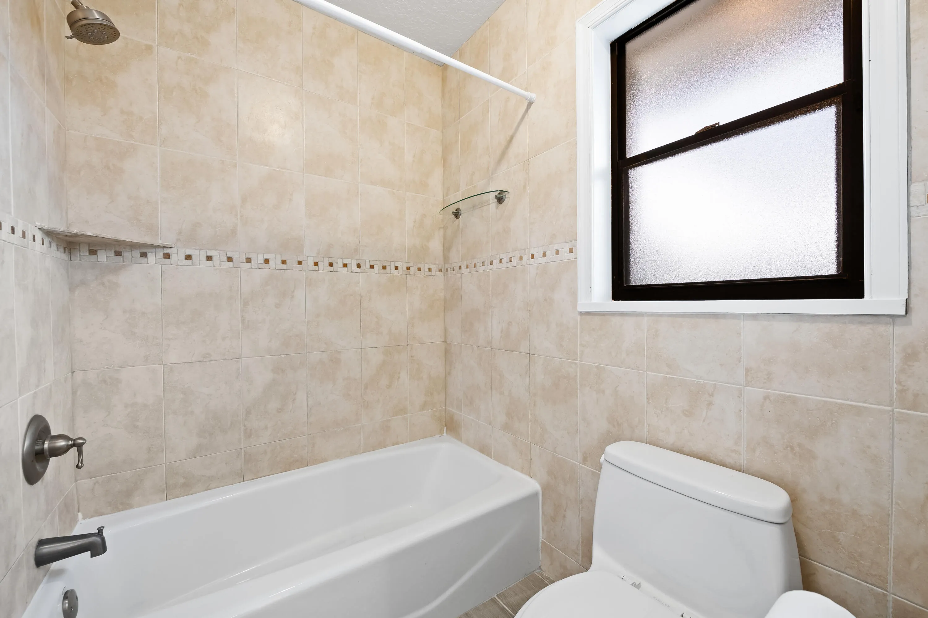 Bathtub with tiled surround and decorative accent band, bronze-finish fixtures, and a privacy window