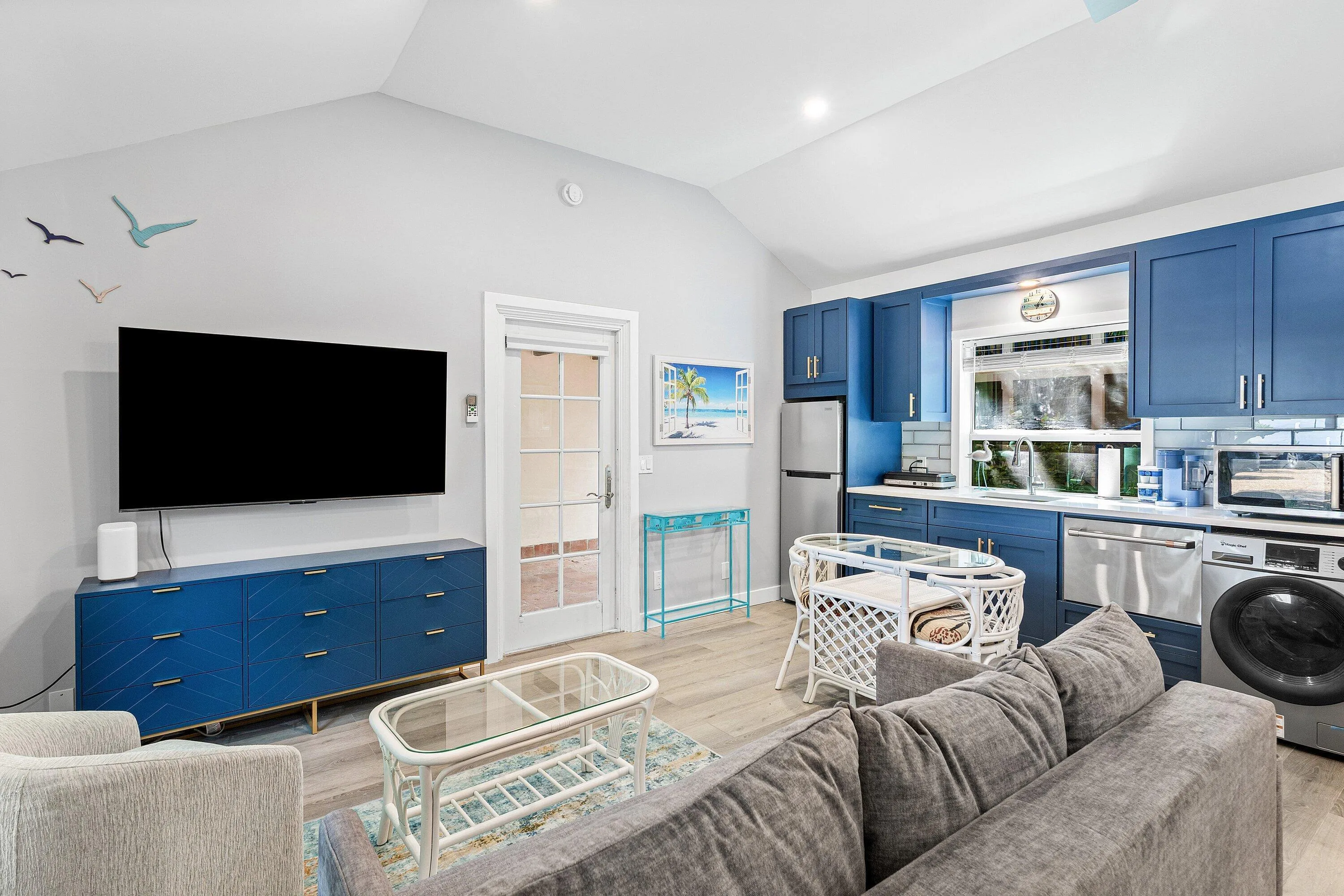 Vaulted ceiling, wood-finish flooring, a kitchen featuring blue cabinetry, stainless steel appliances, and a tile backsplash, and a glass-paned door
