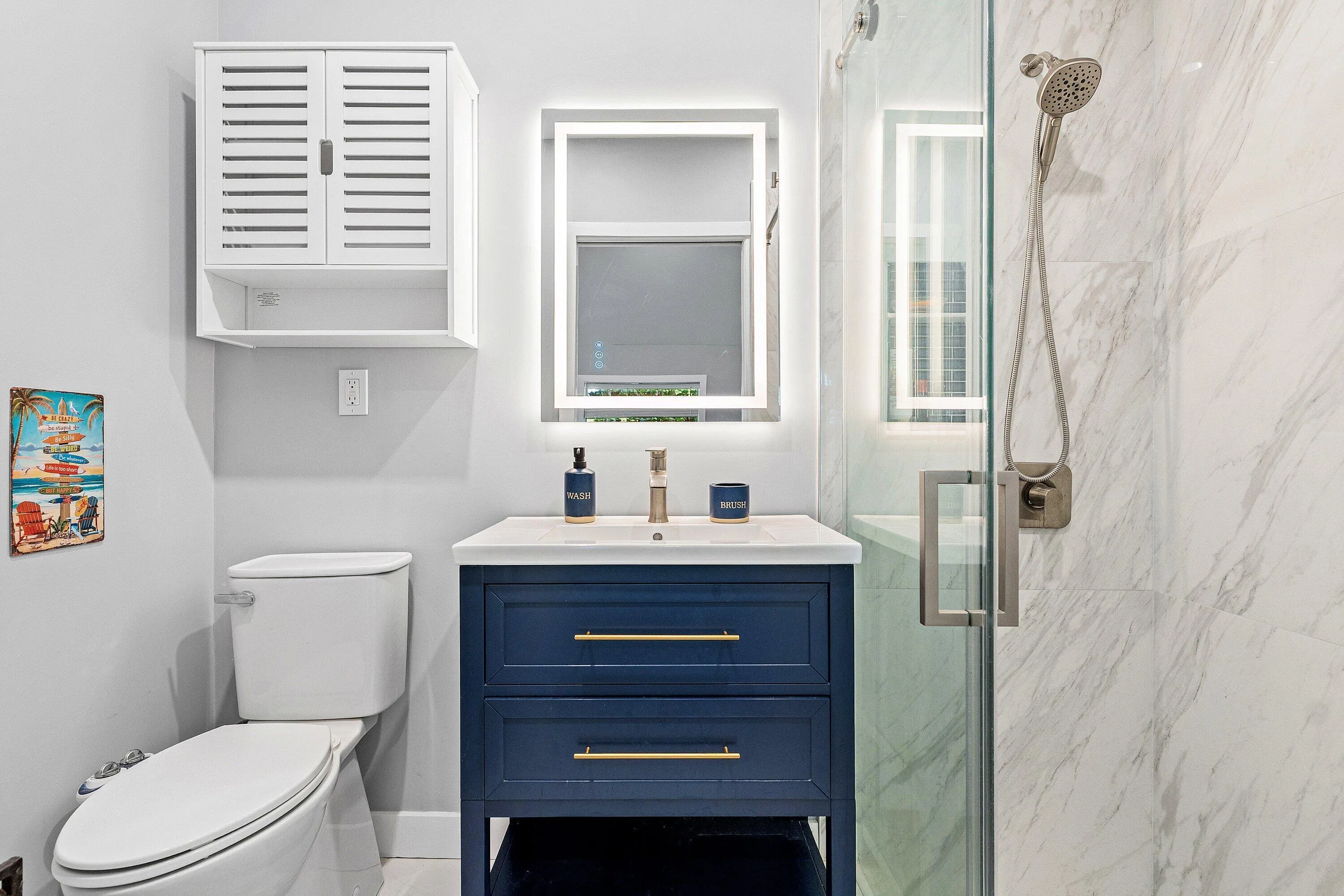 Modern bathroom featuring a blue vanity with gold-finish hardware, illuminated mirror, white countertop, and glass-enclosed shower with marble-style wall tile