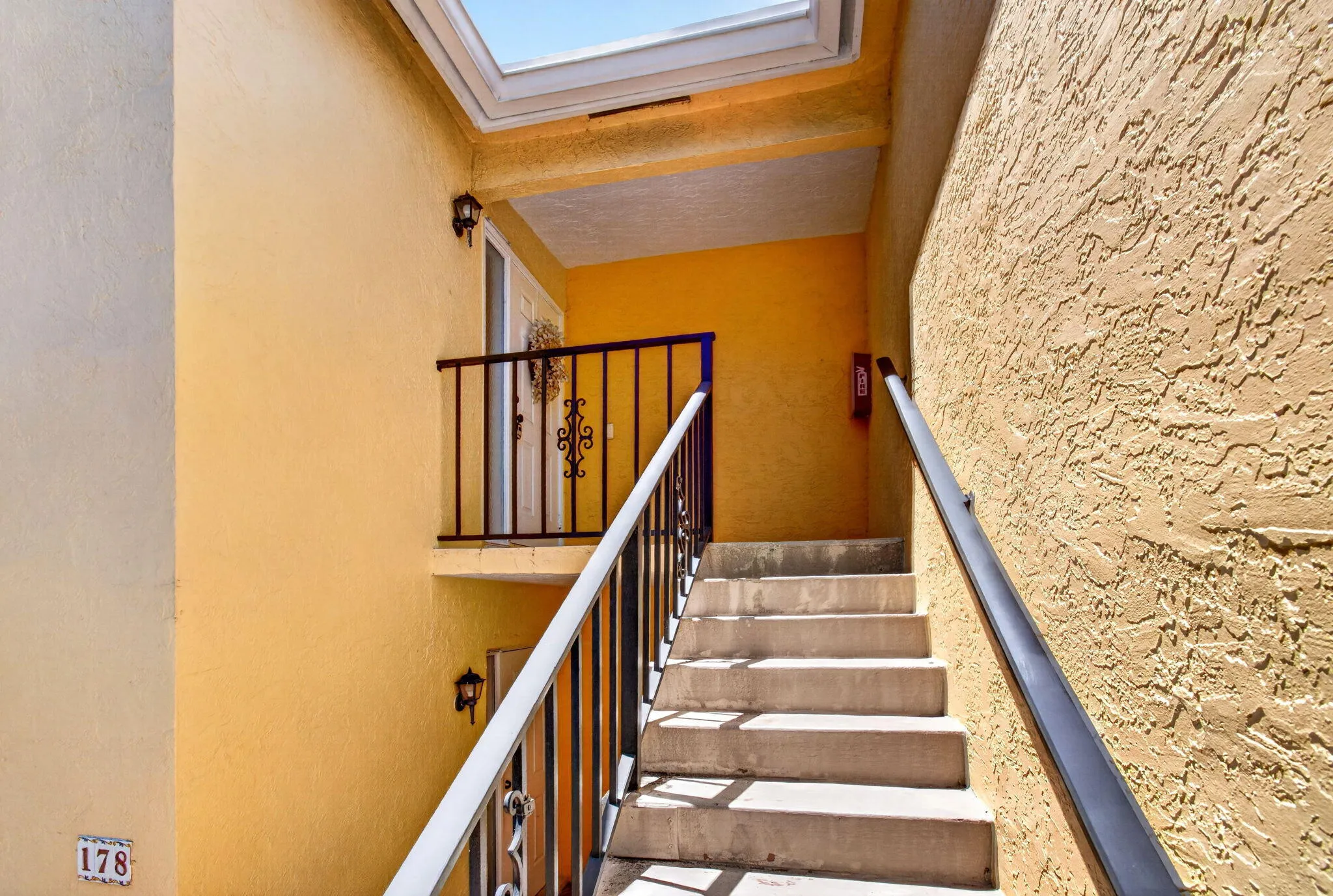 Concrete staircase with black metal railings, textured stucco exterior, exterior wall sconces, and an overhead skylight