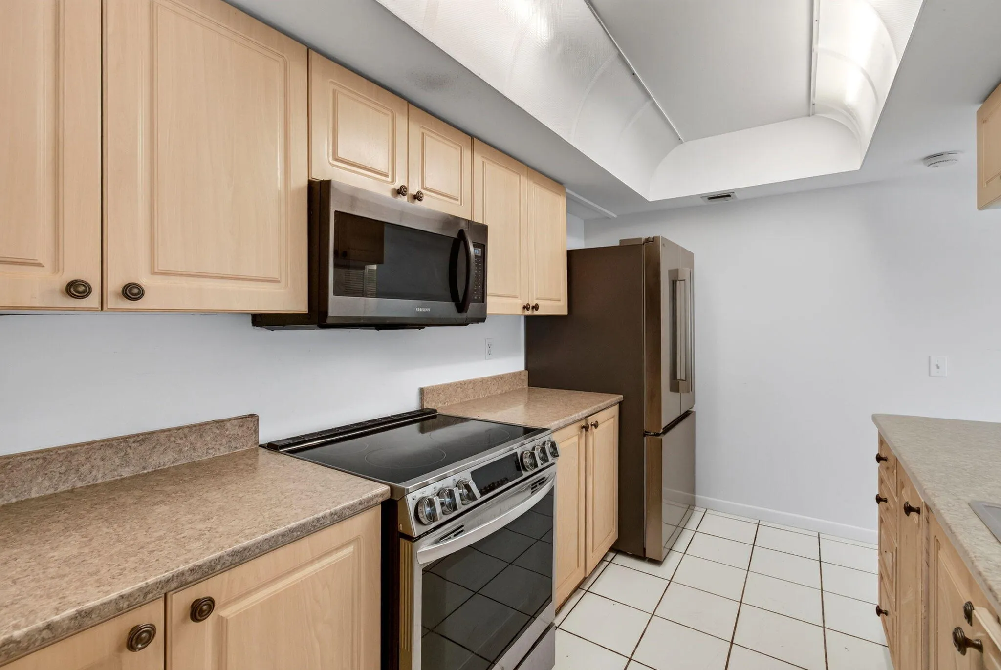 Light wood-finish cabinetry with decorative hardware, stainless steel range and microwave, and white tile flooring