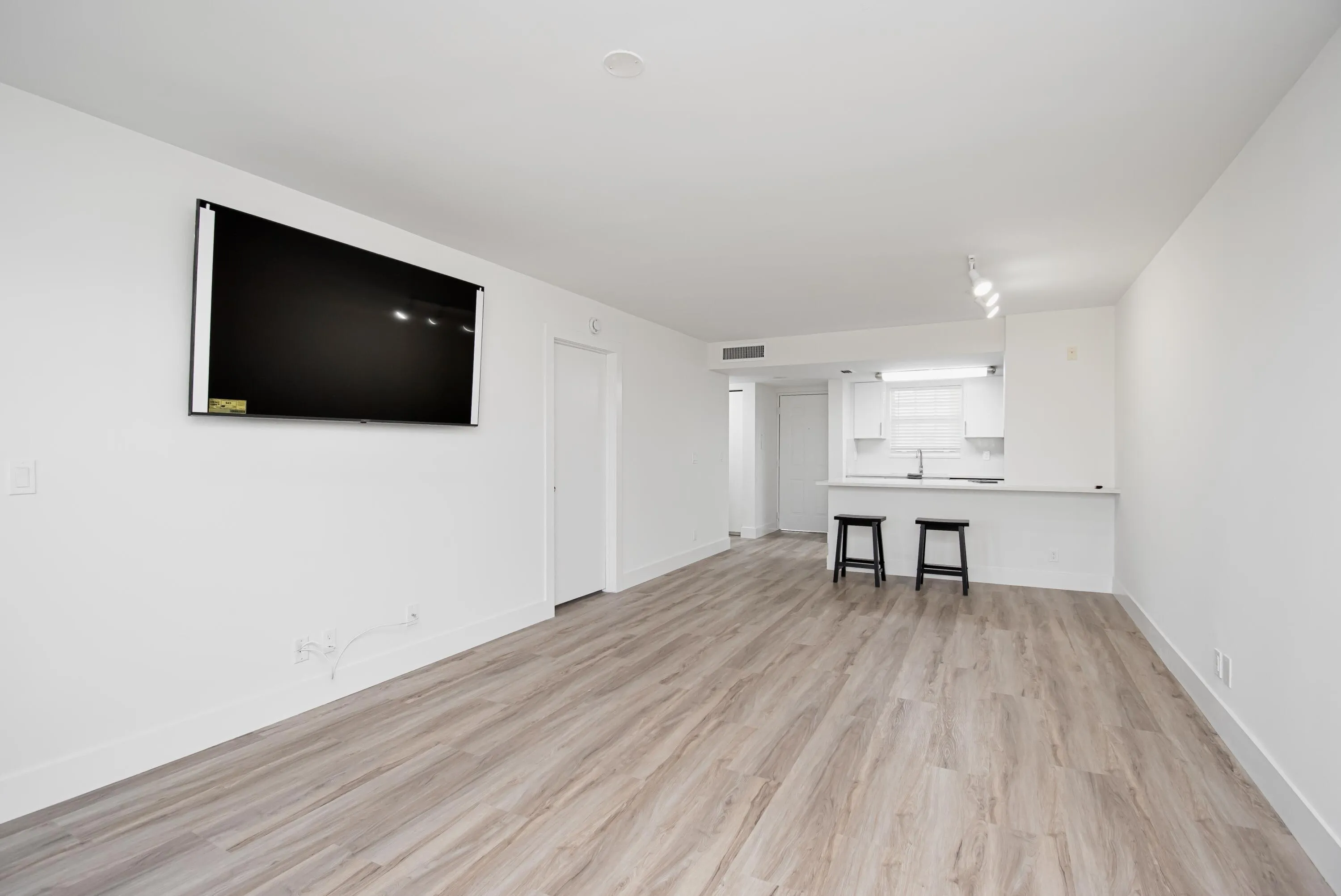 Open-concept living area featuring 65" UHD TV, wood-finish flooring, white walls, and track lighting in the kitchen area