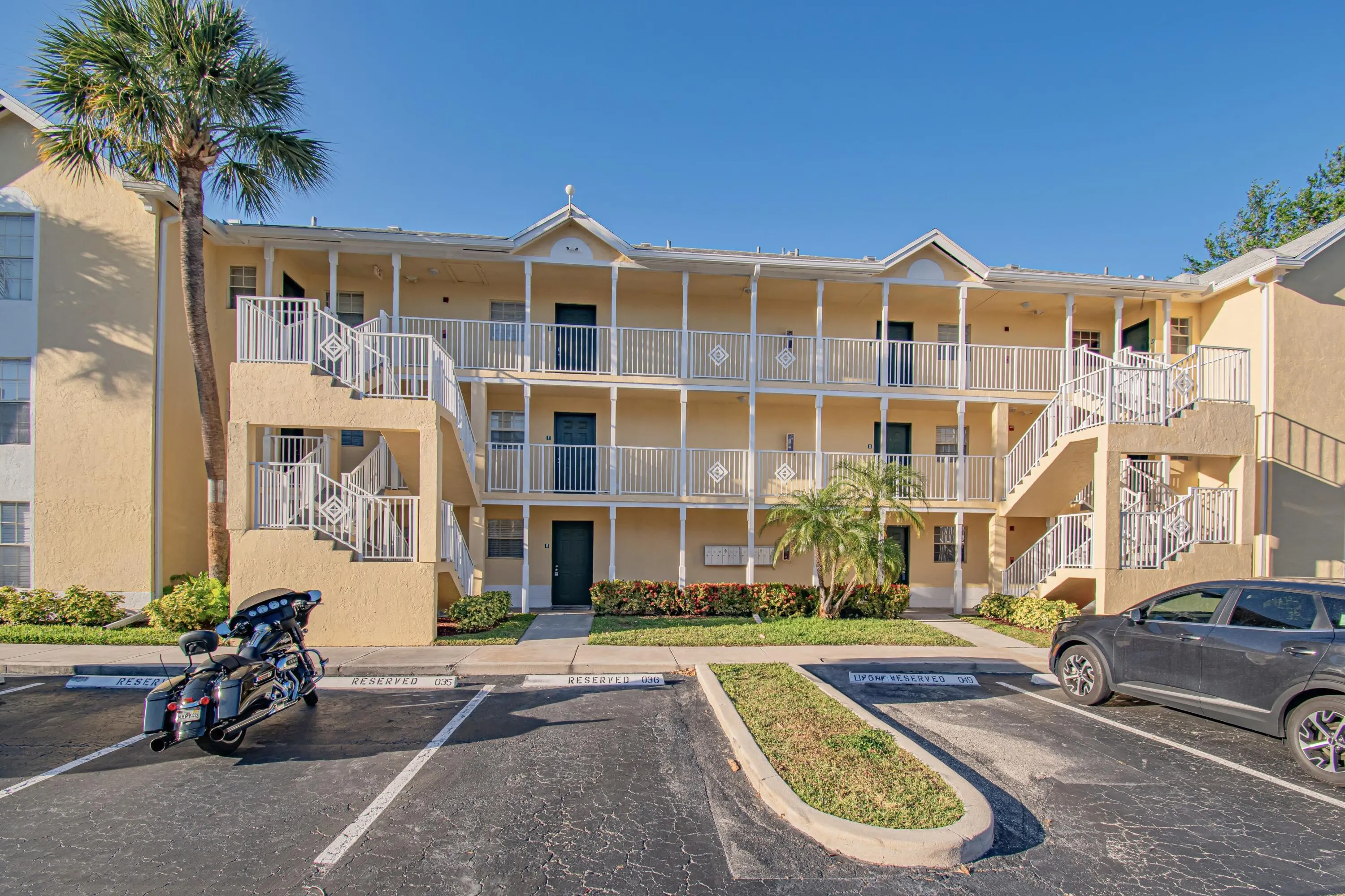 Three-story residential building with stucco exterior, multiple private balconies with white railings, exterior staircases, and mature landscaping
