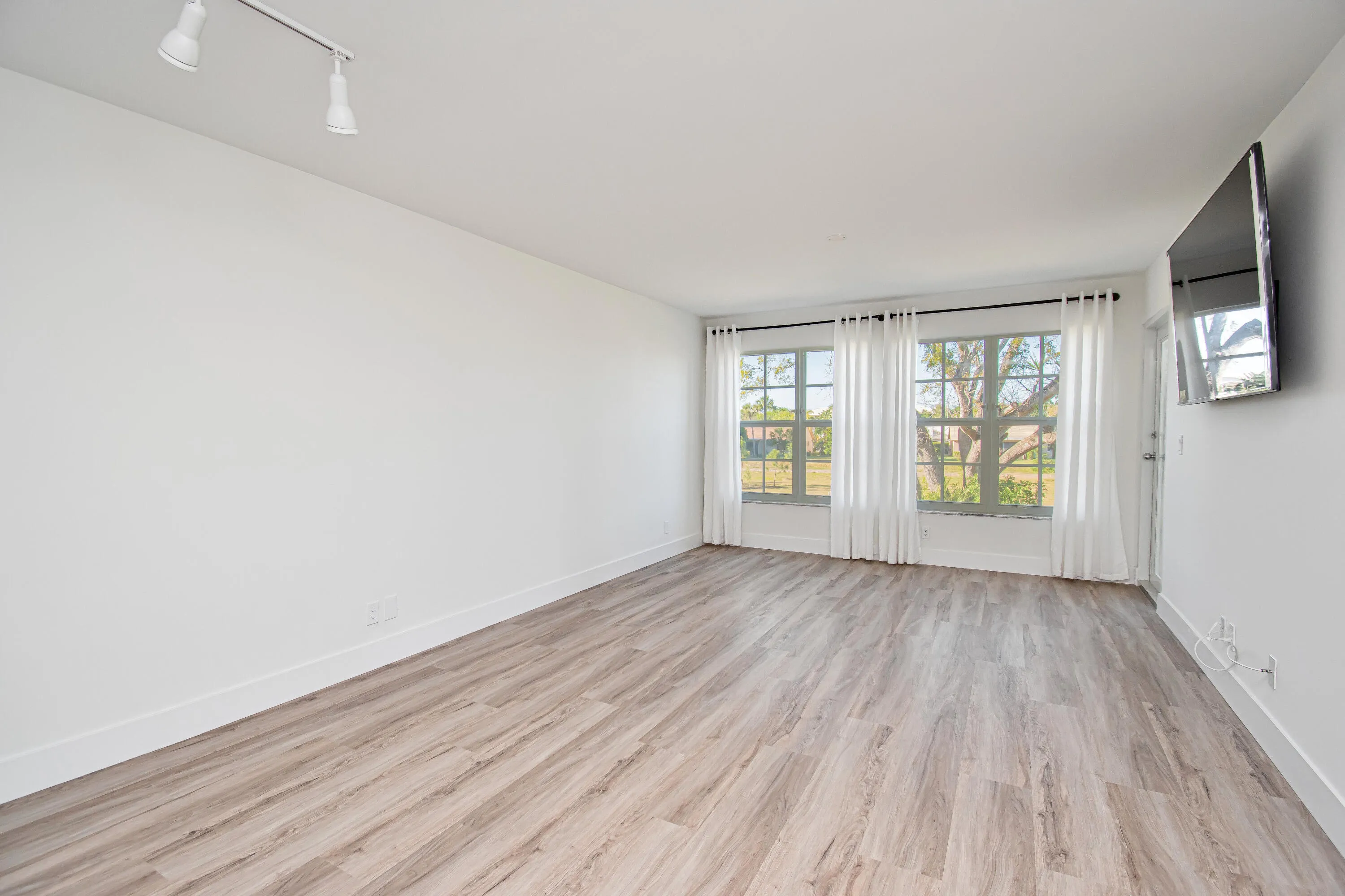 Spacious room featuring wood-finish flooring, white walls, multiple windows with white window treatments, and track lighting