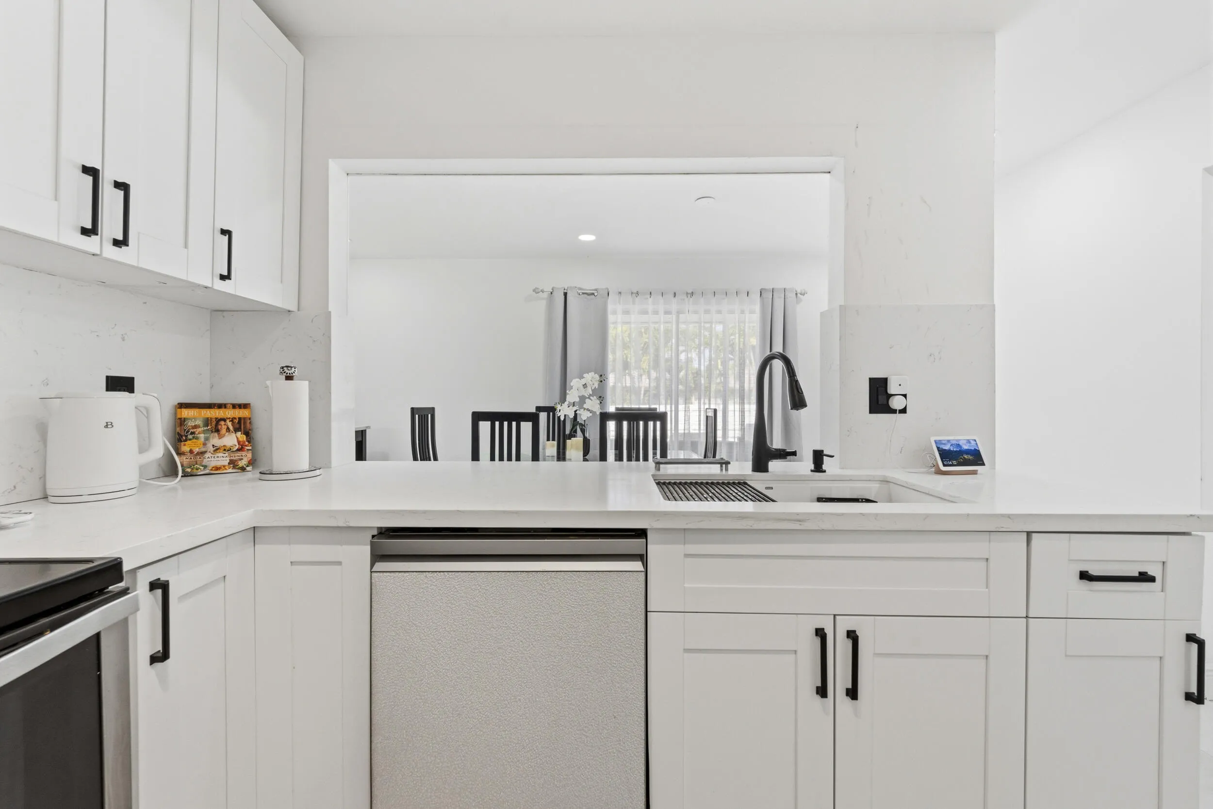 White cabinetry with matte black hardware, light-toned countertops with integrated backsplash, stainless steel sink with a matte black faucet, and a pass-through window to an adjacent room