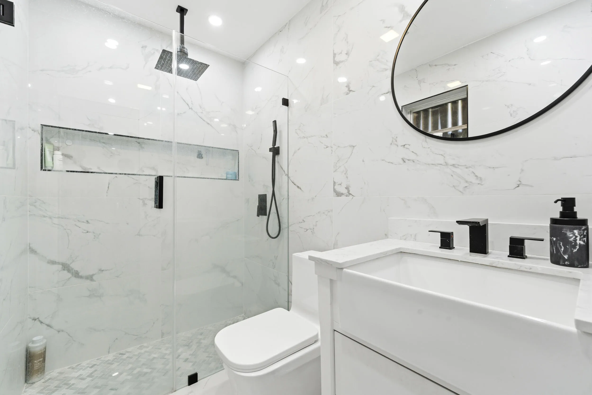Floor-to-ceiling marbled tile, glass-enclosed shower with contrasting matte black fixtures, recessed shower niche, white vanity with integrated sink, and an elegant circular mirror