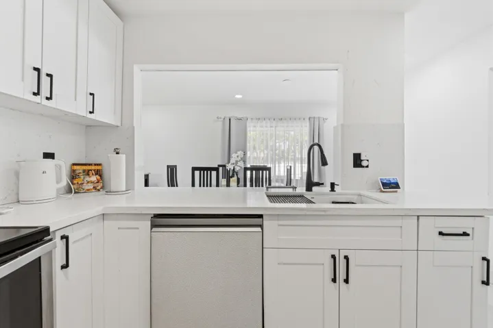 White cabinetry with matte black hardware, light-toned countertops with integrated backsplash, stainless steel sink with a matte black faucet, and a pass-through window to an adjacent room