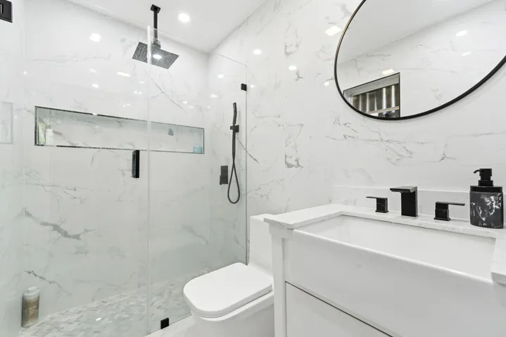 Floor-to-ceiling marbled tile, glass-enclosed shower with contrasting matte black fixtures, recessed shower niche, white vanity with integrated sink, and an elegant circular mirror